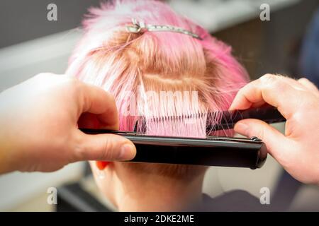 Close up back view of hairdresser's hands straightening short pink hair with a hair iron straightener in a hair salon Stock Photo