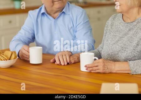 Happy senior couple holding hands while sitting at kitchen table and enjoying tea Stock Photo