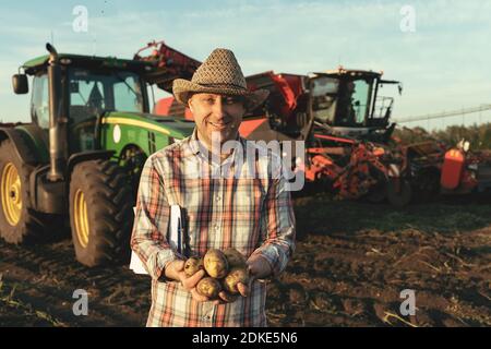 A cheerful farmer on the background of agricultural machinery with potato tubers in his hands. Rich harvest concept. Stock Photo