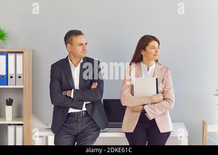 Middle aged man and woman business partners sitting on desk with documents looking aside Stock Photo