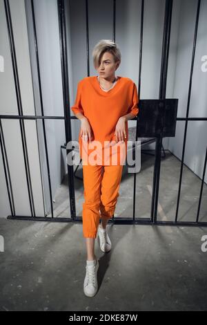 The young criminal is leaning against the bars of the cell in an orange robe. Women's maximum security prison. Stock Photo