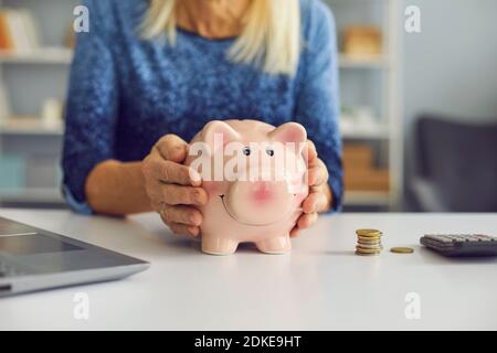Close up of a piggy bank in the hands of an unknown senior woman sitting at a table. Stock Photo