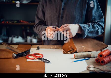 Sitting at a table in the workshop a young guy manually sews leather elements to each other. Stock Photo
