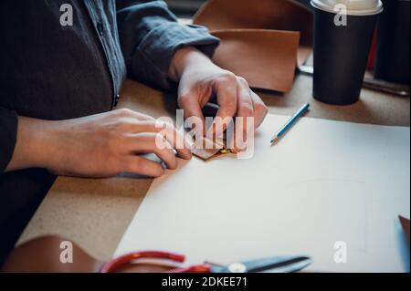 Tailor's hands make loops for the fastener of a women's bag made of genuine leather. Workbench in the workshop. Stock Photo