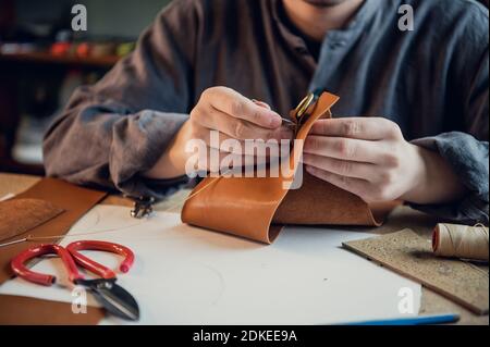 A young experienced shoemaker or tailor sews a unique piece of leather in a spacious bright studio. Stock Photo
