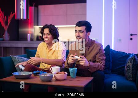 Smiling diverse male friends with gamepads playing video game at home. Portrait of latin and caucasian hipster guys playing game with console relaxing Stock Photo