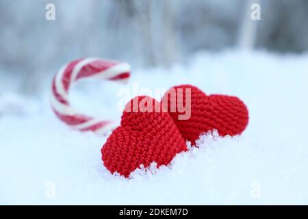 Two love hearts and Christmas candy cane on a snow in winter forest. Red knitted symbols of Valentine's day, background for New Year holiday Stock Photo
