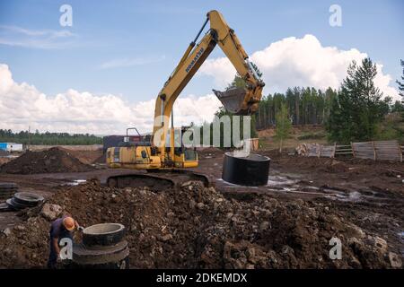 A worker in a pit and an excavator is carrying a concrete ring to install a sewer well on a construction site. Stock Photo