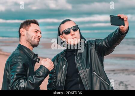 Two Handsome Male Friends Making Selfie Using Black Smartphone at the Beach. Youthful Men in Black Clothes Having Fun by Making Photos Against the Sea Stock Photo