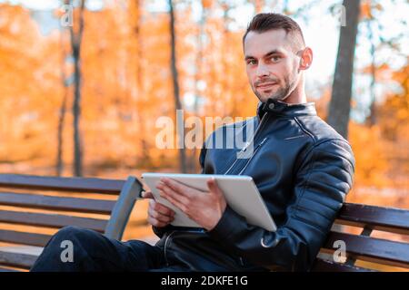 Handsome Young Man Sitting on the Bench and Using Big White Tablet PC at the Beautiful Autumn Park Stock Photo