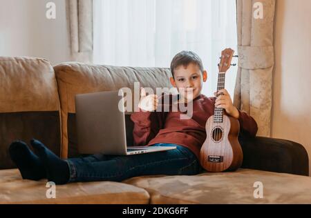 Boy playing ukulele and watching online course on laptop while practicing at home. Online training, online classes.Vintage color filter Stock Photo