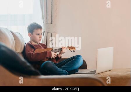 Boy playing ukulele and watching online course on laptop while practicing at home. Online training, online classes.Vintage color filter Stock Photo