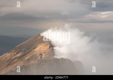 Epic view of mountain hiker standing on the top of the cliff and distant mountain ridge lighten by soft sunlight from one side and covered by fog Stock Photo