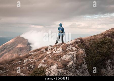 Young mountain hiker with blue jacket and backpack standing at the edge of cliff looking at the distant, rocky, misty ridge under cloudy, stormy sky Stock Photo