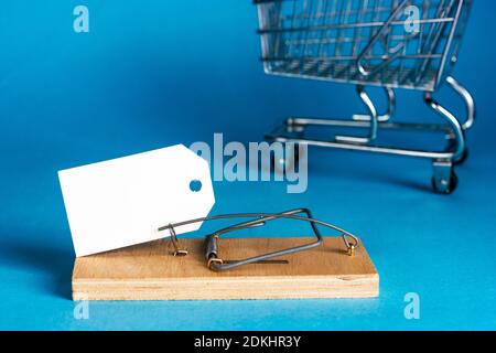 White paper plate in a wooden mousetrap on a blue background. Supermarket cart in the background. Marketing gimmicks. Consumer deception. Place for Stock Photo