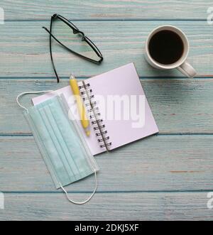 Open notepad with face mask, pen, spectacles and coffee on blue wood table. Top view. Stock Photo