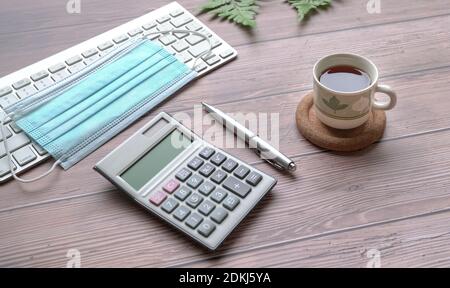 Wood table with keyboard, face mask, black coffee, pen, calculator and green plants.