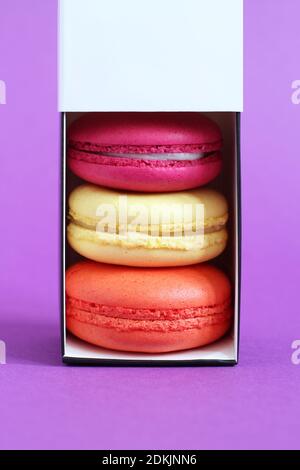 Multicolored macarons in a cardboard box with an open lid standing upright on a lilac background. Closeup Stock Photo