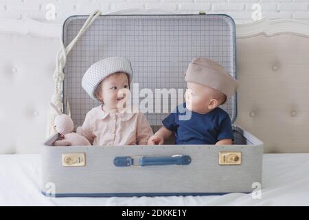Cute Girl And Boy Sitting In Suitcase On Bed At Home