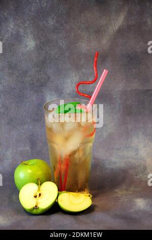 A tall glass of a pale yellow drink with ice and straws on a gray background next to ripe green apples. Vertical positioning. Stock Photo
