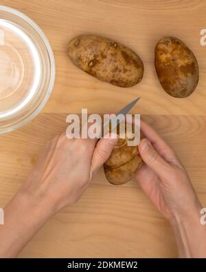 Woman hands peeling potatoes, close up view from above, wooden rustic background, vertical banner Stock Photo
