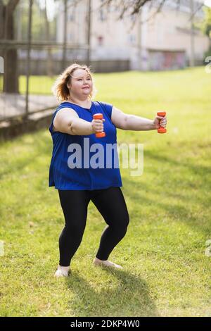 A young plus size blonde performs an exercise with dumbbells outdoors. Healthy lifestyle concept. Stock Photo