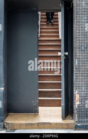 an open grey door with surrounding tile, with a sodden staircase visible and the legs of a person walking down Stock Photo