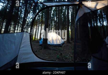 Evening camping in the forest. Silhouette of two tourists sitting in white tent, enjoying sunset. View from inside a tent. Tourism adventure active lifestyle concept Stock Photo