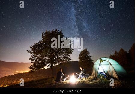 Young couple travellers resting at campfire near tourist tent, warming hands under amazing night sky full of stars and Milky way. On the background starry sky, mountains, big tree and luminous town Stock Photo