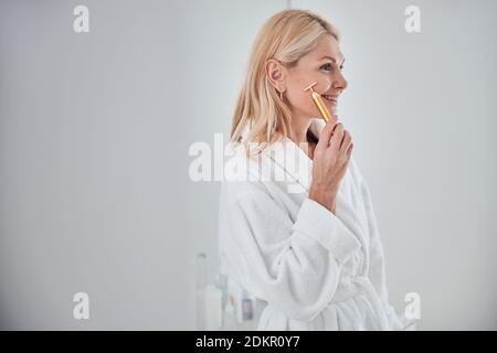 Adult female in white plush bathrobe holding face massager in hand isolated on white background Stock Photo