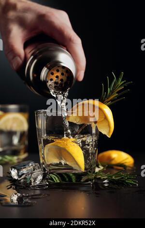 Cocktail Gin and Tonic with lemon and rosemary. The bartender pours a cocktail from a shaker into a glass. Stock Photo