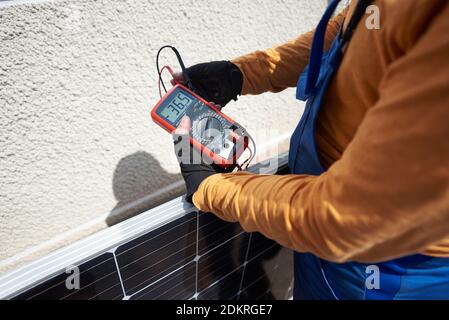 Male worker installing solar photovoltaic panel system on roof of modern house. Professional electrician checking blue solar module, using voltmeter. Alternative energy ecological concept. Stock Photo