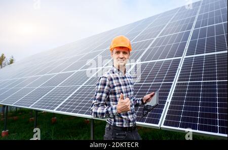 Electrician in a helmet carries out work in a tablet on the background of a large plantation of solar panels. Photovoltaic power supply systems. Green energy concept design. Stock Photo