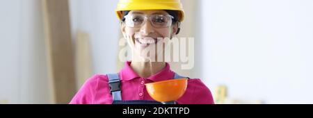 Smiling woman in yellow hard hat and goggles holding plunger in hand Stock Photo