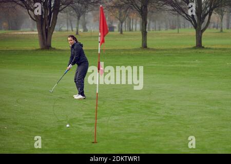 Middle-aged fit woman golfer putting for the hole on the green viewed from the flag as her golf ball approaches on a cold windy misty day Stock Photo