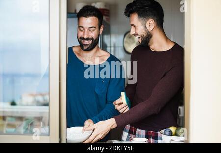Window view of gay couple washing dishes together inside home kitchen - Young hosexual people having fun during morning routine - Focus on right man f Stock Photo