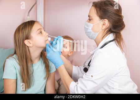 Pediatrician taking nasal mucus test sample from elementary age girl's nose performing respiratory virus testing procedure at home during coronavirus Stock Photo