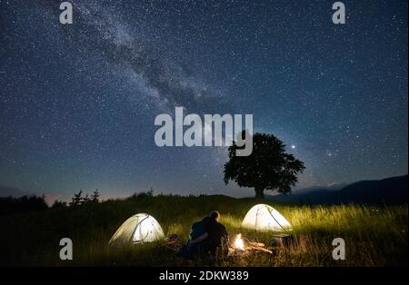 Friends sitting on a bench made of logs, watching fire together hugging between two illuminated tents, silhouette of a big tree on background, Milky Way and starry sky above them, starry night concept Stock Photo