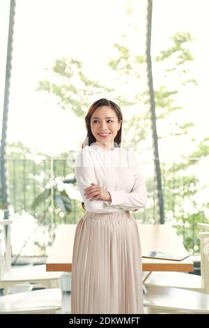 Happy young beautiful Asian woman with brace retainers, standing arms crossed, smiling to camera in modern office with big glass window and blur green Stock Photo