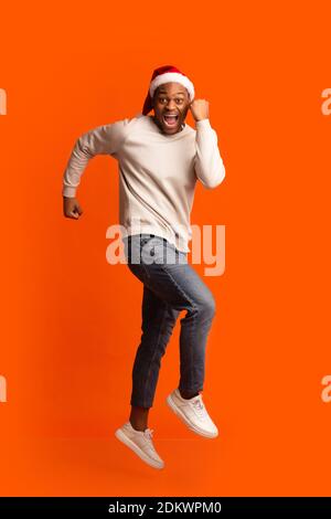 Ready For Party. Cheerful African Guy In Santa Hat Running In Air Stock Photo