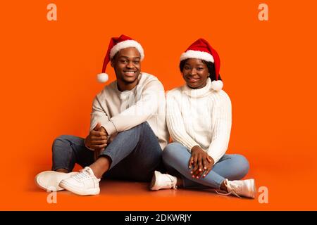Merry Christmas. Smiling Black Couple In Santa Hats Sitting Smiling At Camera Stock Photo