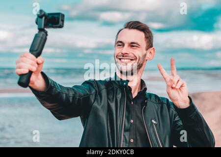 Smiling Young Blogger Making Selfie or Streaming Video at the Beach Using Action Camera with Gimbal Camera Stabilizer. Man in Black Clothes Making Photo Showing Peace Stock Photo