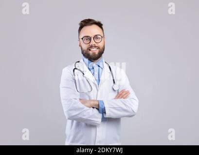 Portrait of happy young doctor in lab coat standing with crossed arms and looking at camera over grey background Stock Photo