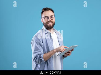 Positive young guy in casual wear using tablet computer over blue studio background Stock Photo