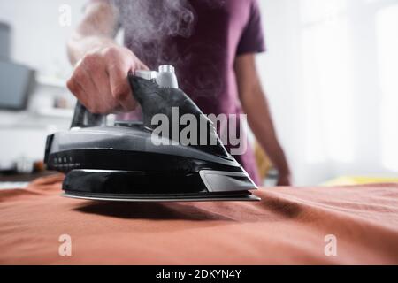 Cropped view of iron with steam near clothes in hand of man on blurred background Stock Photo