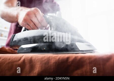 Cropped view of iron with steam on clothes in hand of man on blurred background Stock Photo