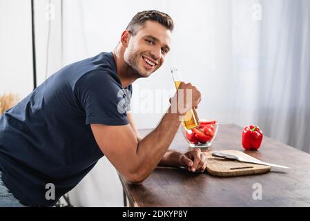 Side view of smiling man holding bottle of beer near cutting board and bell pepper on blurred background Stock Photo