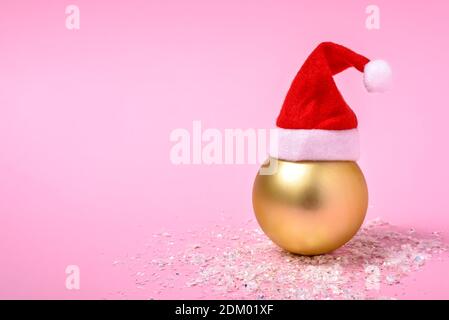 Golden Christmas bauble with Santa Claus hat on pink background with copy space for text. Creative Christmas and New Year concept. Stock Photo