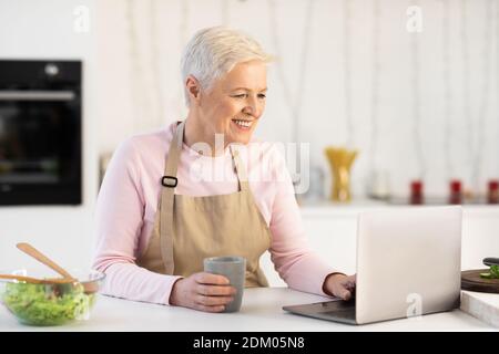 Happy Senior Woman Using Laptop Watching Cooking Video In Kitchen Stock Photo