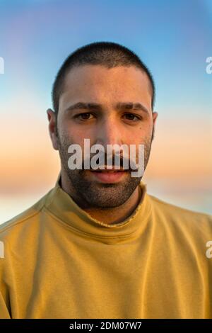 Portrait of young man with moustache looking into camera in front of sunset sky wearing yellow jumper Stock Photo